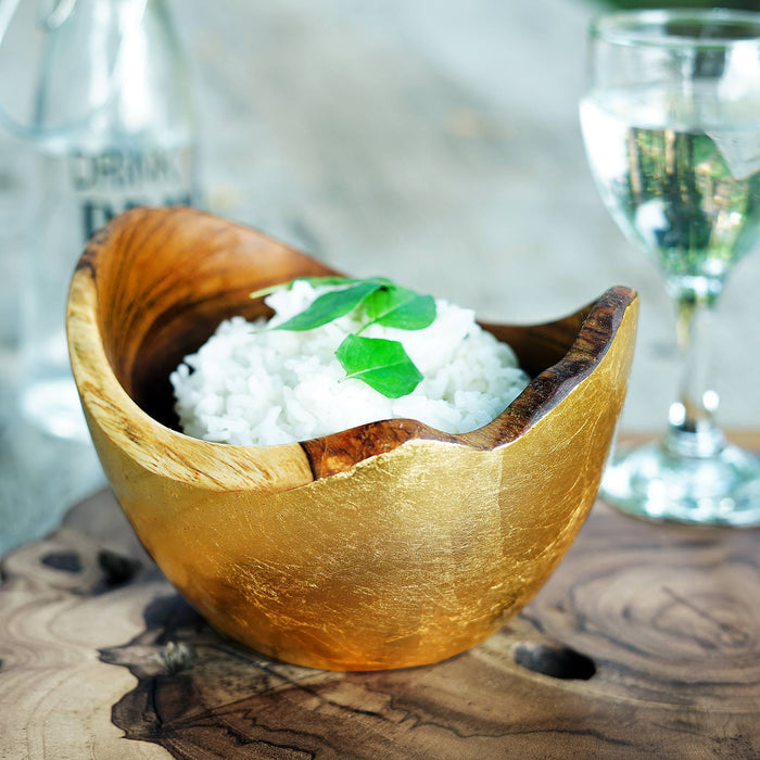 Rico & Plato adagio teak bowl with gold leaf exterior with rice inside on a wooden surface with glasses of water in the background