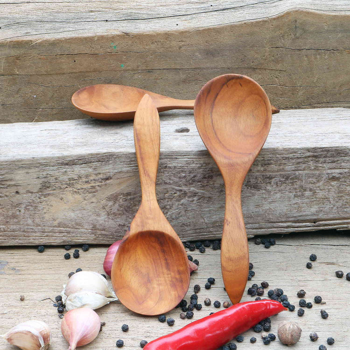 A set of three wooden spoons with a golden-brown hue, displayed on a wooden surface surrounded by spices, garlic, and a red chili.
