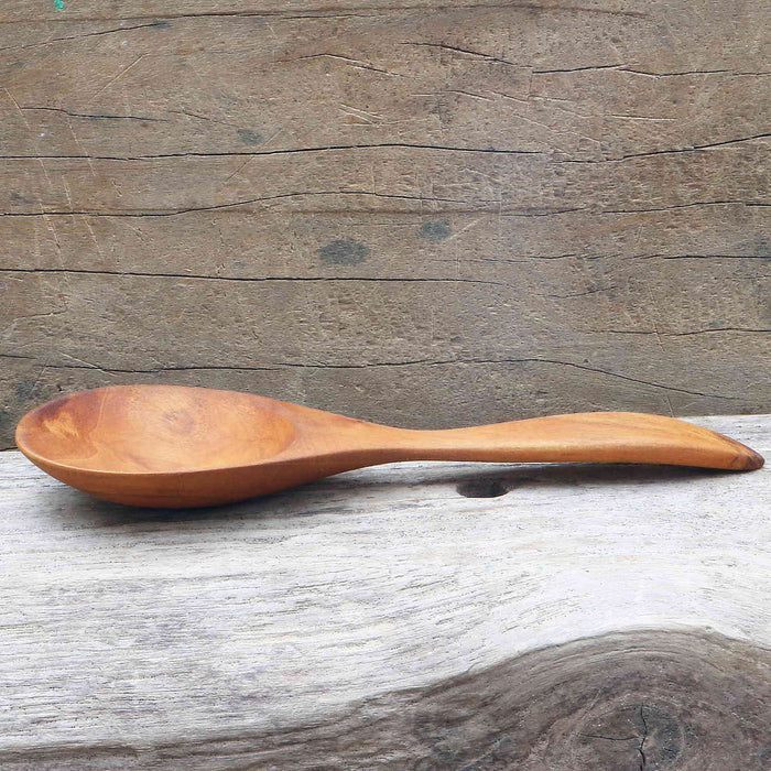 Side view of Rico & Plato almira teak spoon on a wooden surface with a wooden background