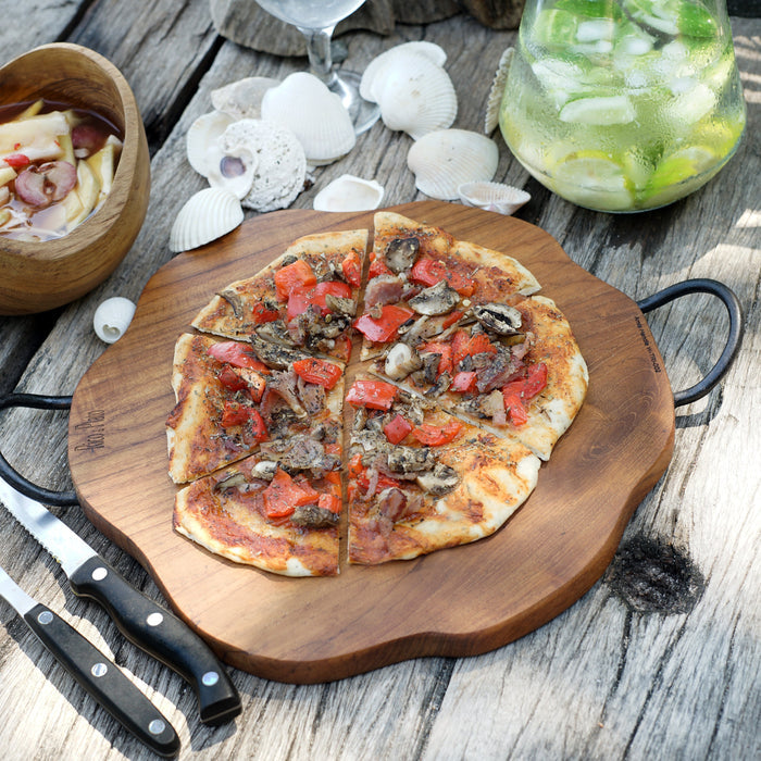 Pizza board in natural teak with metal handles with a pizza on top next to drinks displayed on a wooden surface