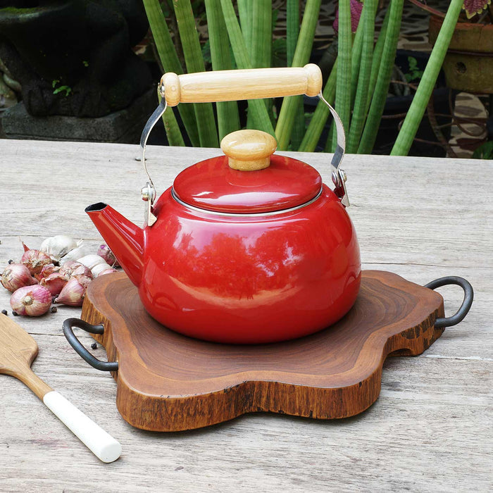 Rico & Plato amla teak trivet with a red kettle on top next to a spatula, shallots, garlic, and peppercorns on a wooden surface outside