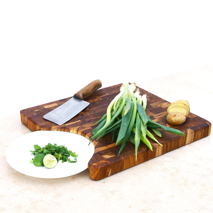 A rectangular, end grain teak cutting board with a cutout for a plate, accompanied by a knife and vegetables on top, displayed on a white surface.