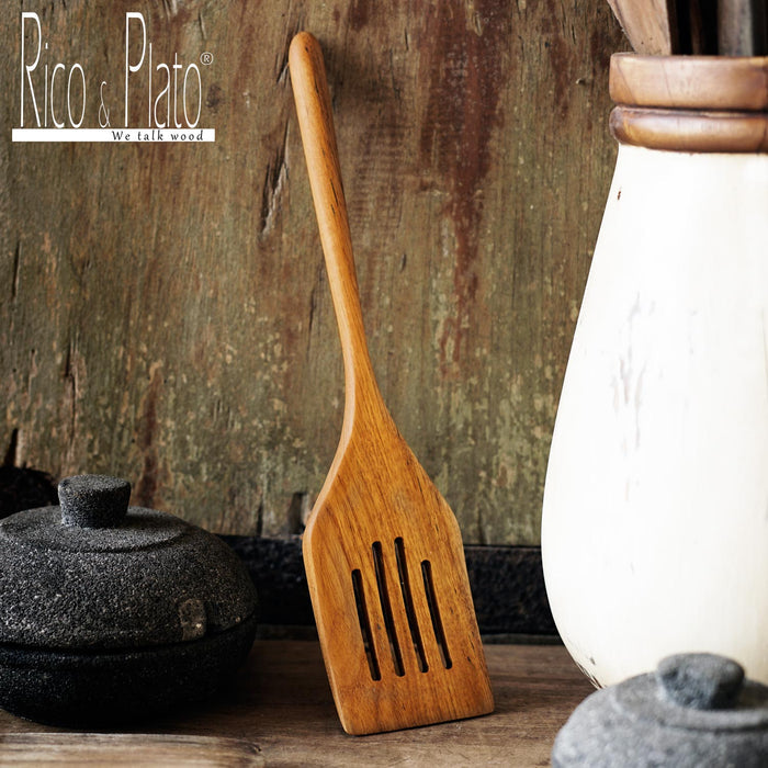 A wooden spatula with drainage slots, resting against a rustic background, part of the kitchen utensils collection by Rico & Plato.