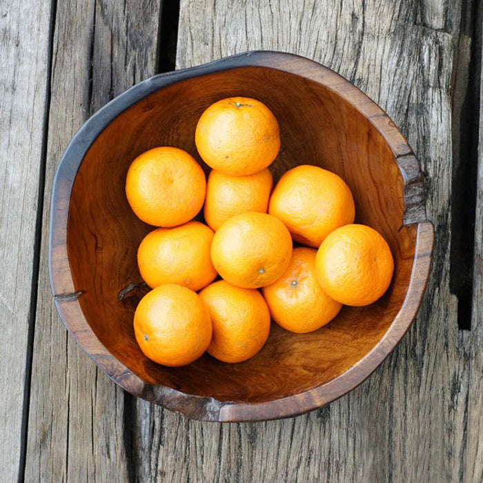 A teak wood bowl with a black burned exterior finish, filled with oranges, placed on a wooden surface.