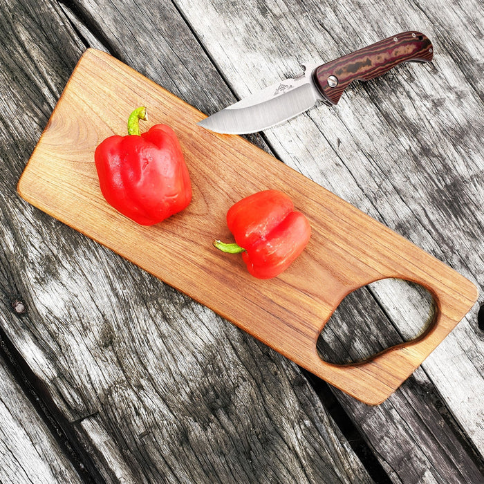Top view of teak cutting board with hole for handle with knife and peppers displayed on a wooden surface