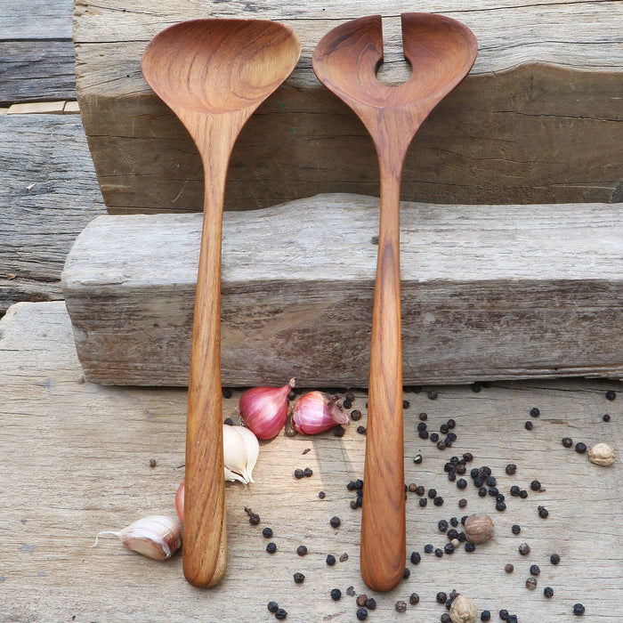 Two handcrafted teak wood salad servers with smooth, ergonomic handles, placed on a wooden surface surrounded by scattered black peppercorns and garlic cloves.