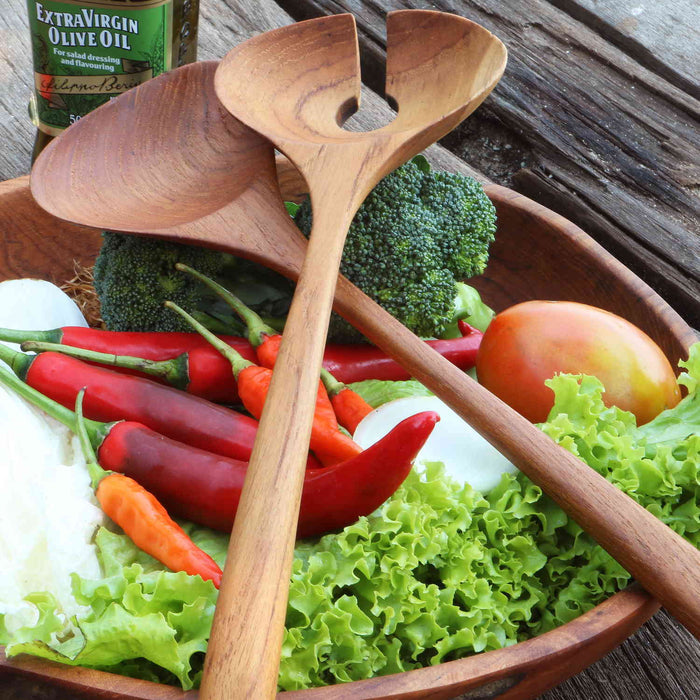 Two teak salad serving utensils placed on a wooden bowl with fruit and veggies on a wooden background
