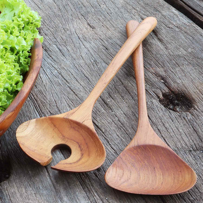 Two teak salad serving utensils on a wooden surface next to lettuce in a wooden bowl