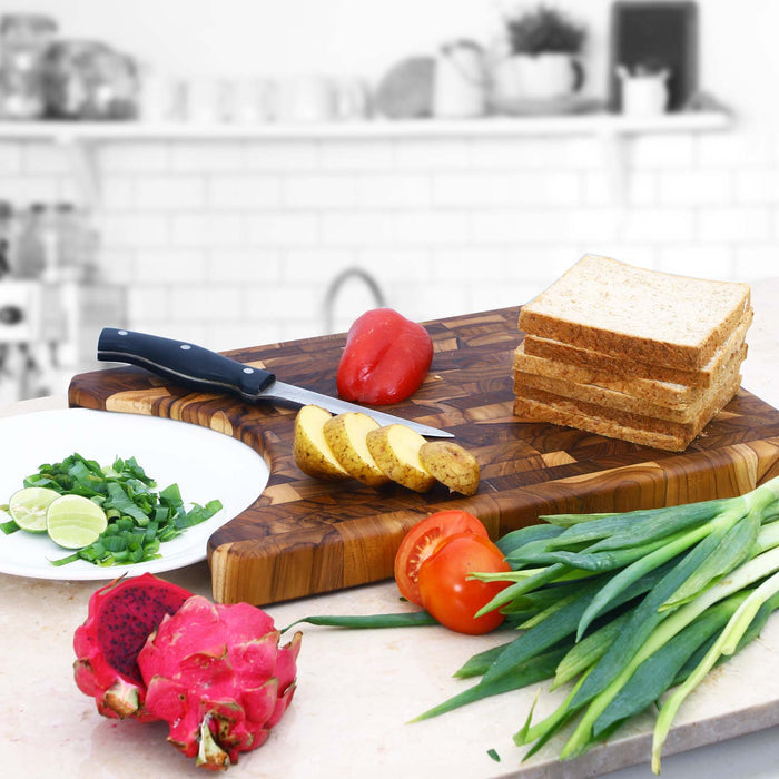 A rectangular end grain teak cutting board with a cutout on the side, placed on a kitchen counter surrounded by various fruits and vegetables such as dragon fruit, tomatoes, and green onions.