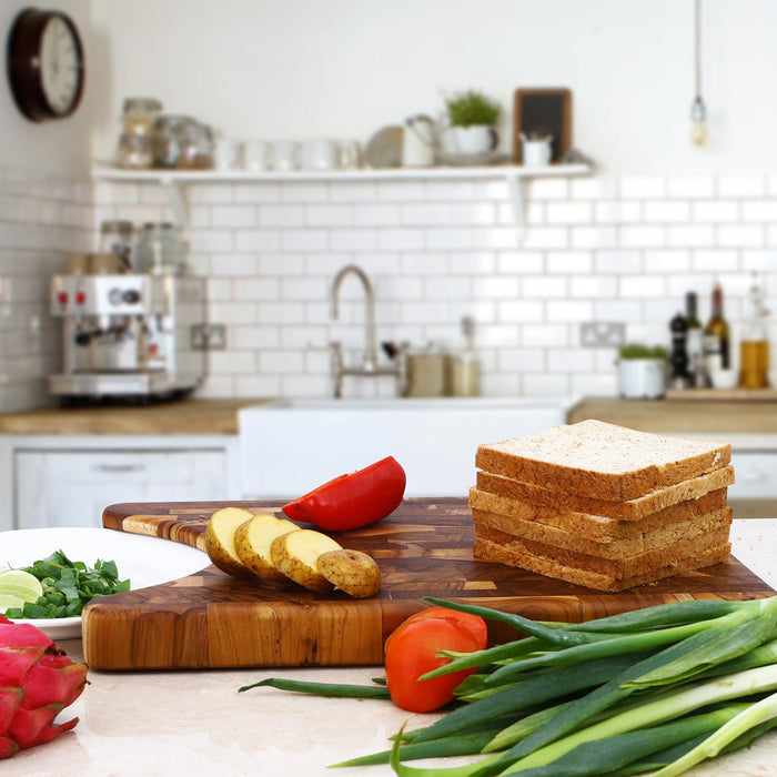 End grain teak cutting board with a plate indent with bread, potatoes, tomatoes, green onions, plate with chopped food displayed on top in a kitchen setting