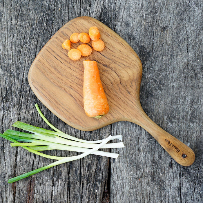 A wooden cutting board with a sleek, rounded edge profile and an ergonomic handle, displayed on a rustic wooden surface with some carrots and green onions beside it.