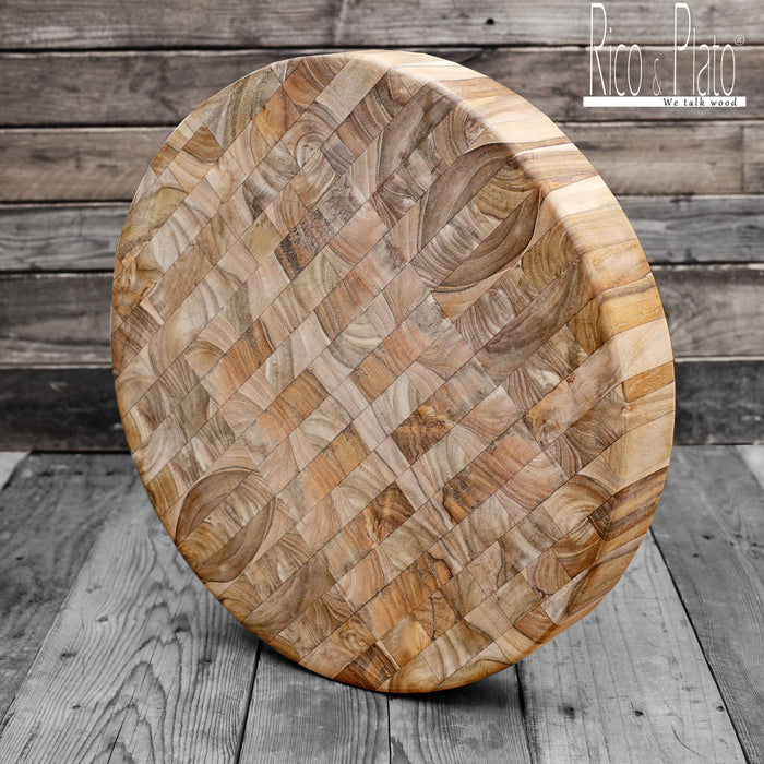 A round, teak wood end grain cutting board with a checkered pattern, displayed on a wooden surface.