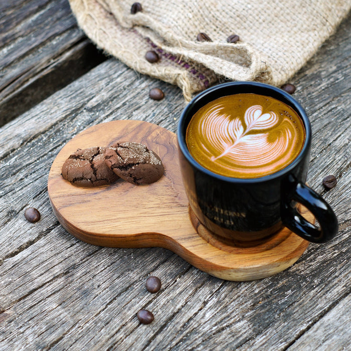 A bean-shaped coffee tray made of teak wood, with a recess to hold a mug, accompanied by a cup of coffee and a cookie. The tray is placed on a wooden surface with coffee beans scattered around.