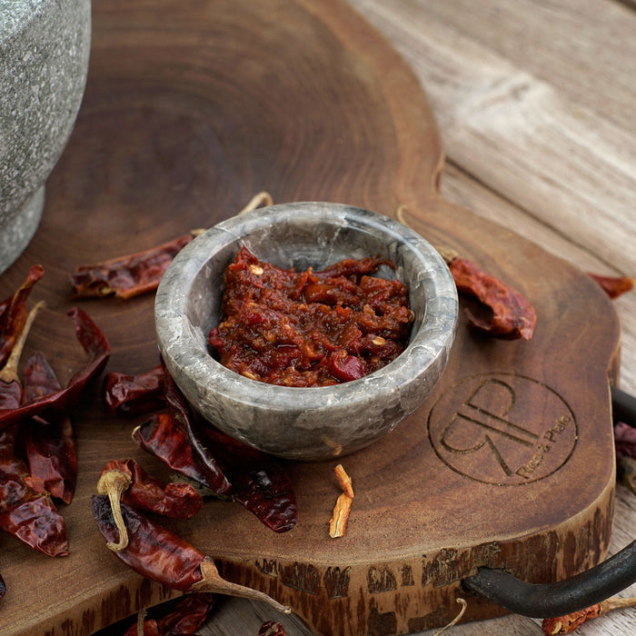 Sambal bowl with sambal sauce inside next to chili peppers on a wooden trivet