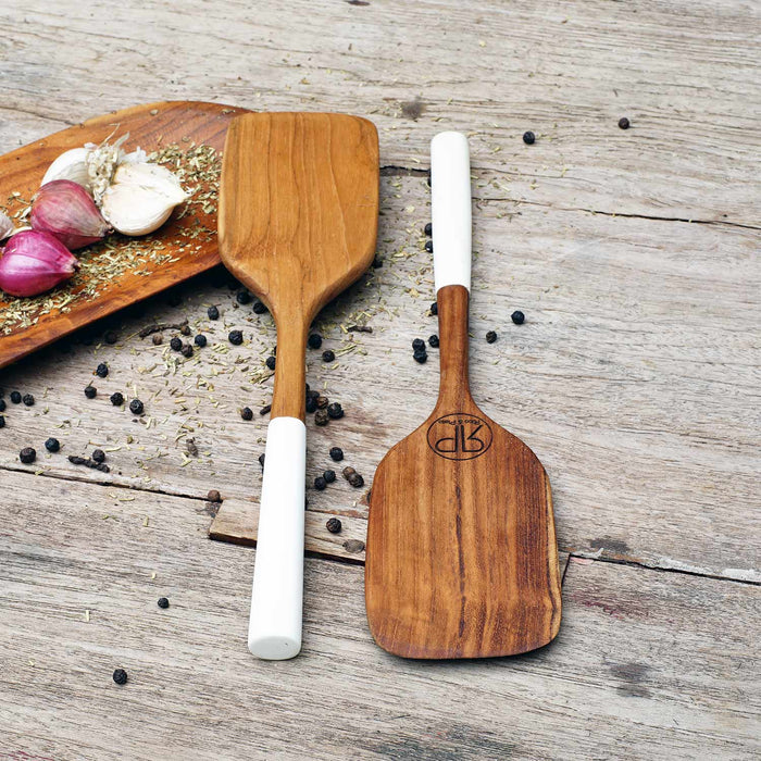 2 Rico & Plato bistro teak spatula with white handles next to a large wooden plate with herbs, garlic, shallots, and peppercorns on a wooden surface