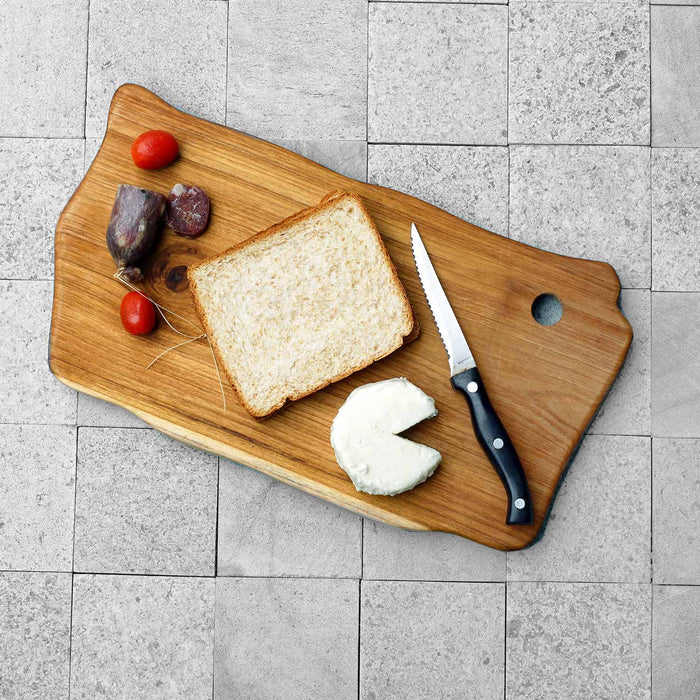 Top view of teak cutting board with meat, tomatoes, cheese, bread and knife displayed on a grey tile surface