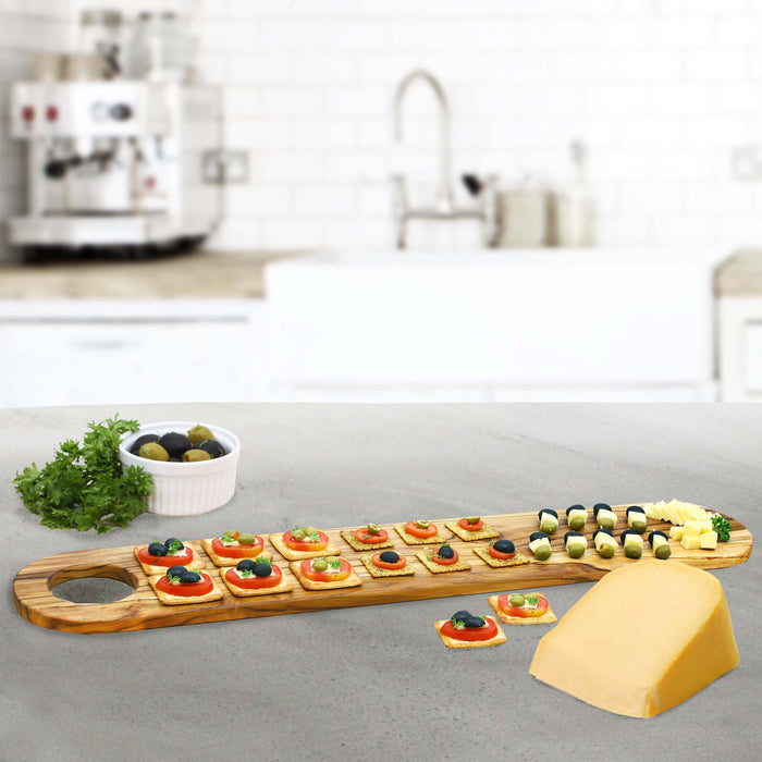 Rico & Plato teak serving board with charcuterie material next to a bowl of olives and cheese in a kitchen setting