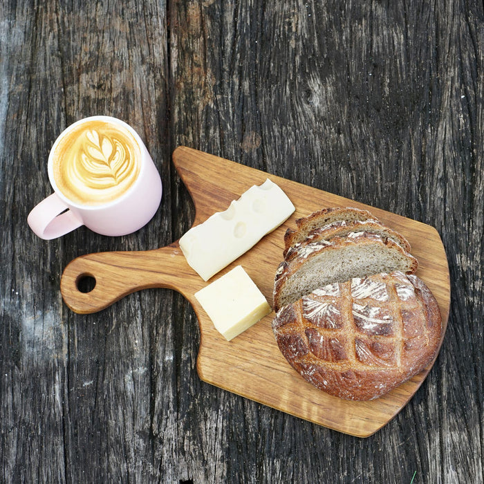 Top view of Rico & Plato teak cutting board/serving platter with cheese and bread on board next to creamy coffee mug displayed on a wooden surface
