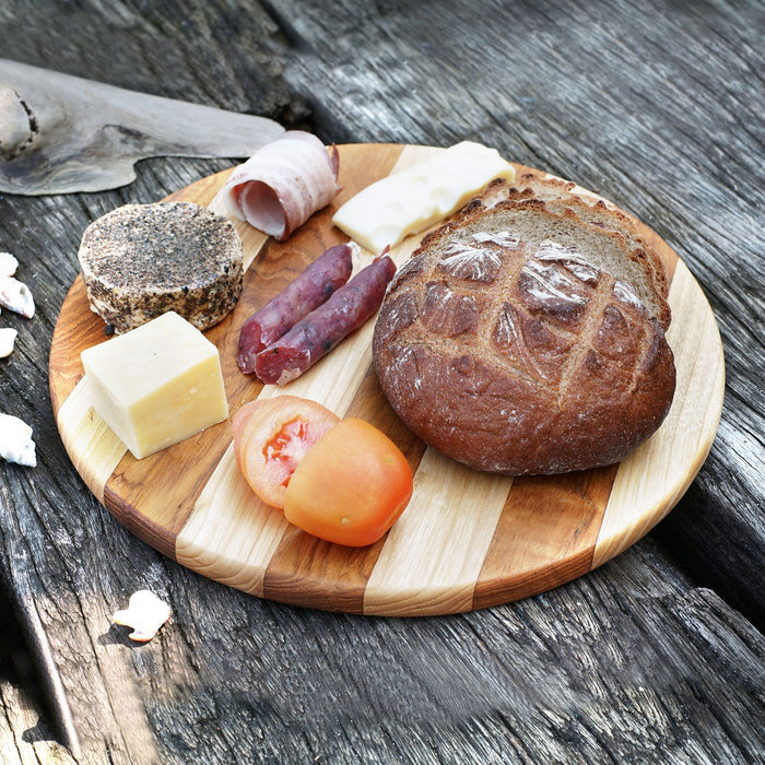Circular teak cutting board/serving platter with 2 tones of wood and meat, cheese, tomatoes, and bread displayed on a wooden surface