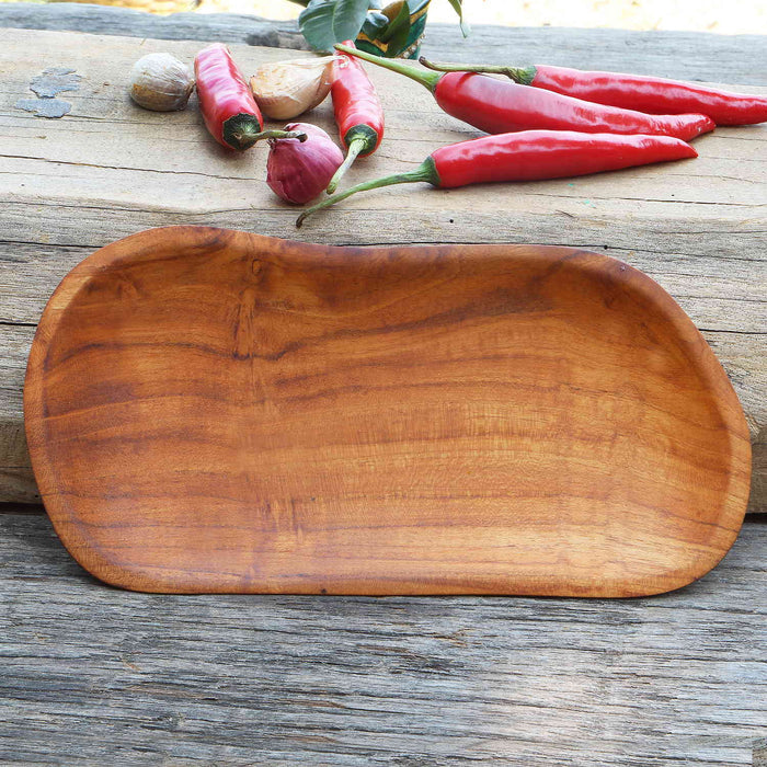 Small teak plate displayed horizontally on a wooden surface next to chili peppers in the background