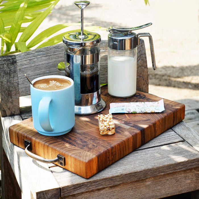 A rectangular, end grain teak cutting board with brass handles, presented as a coffee serving tray. It is displayed outdoors with a cup of coffee, milk, and french press on top.