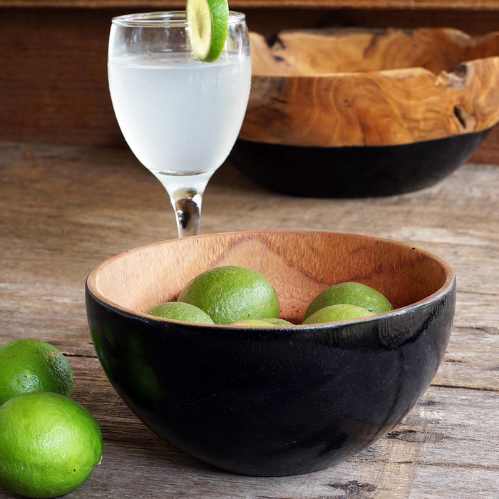 Rico & Plato chico teak bowl with black exterior with limes inside next to 2 limes and a tall wine glass with a clouding white drink and lime slice on the rim all displayed on a wooden surface
