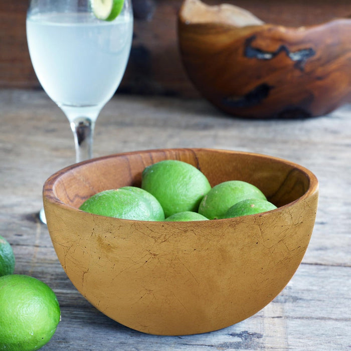 Teak bowl with gold leaf exterior with limes inside and glass of water int he background displayed on a wooden surface