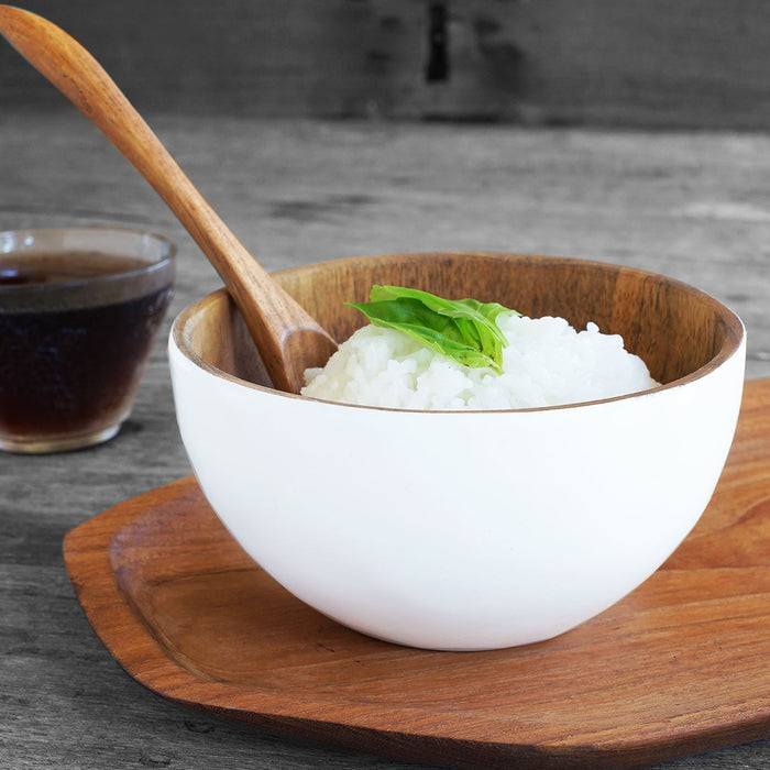 Rico & Plato chico teak bowl with white exterior with rice and a wooden spoon inside on a wooden plate next to a clear glass of brown liquid all on a wooden surface