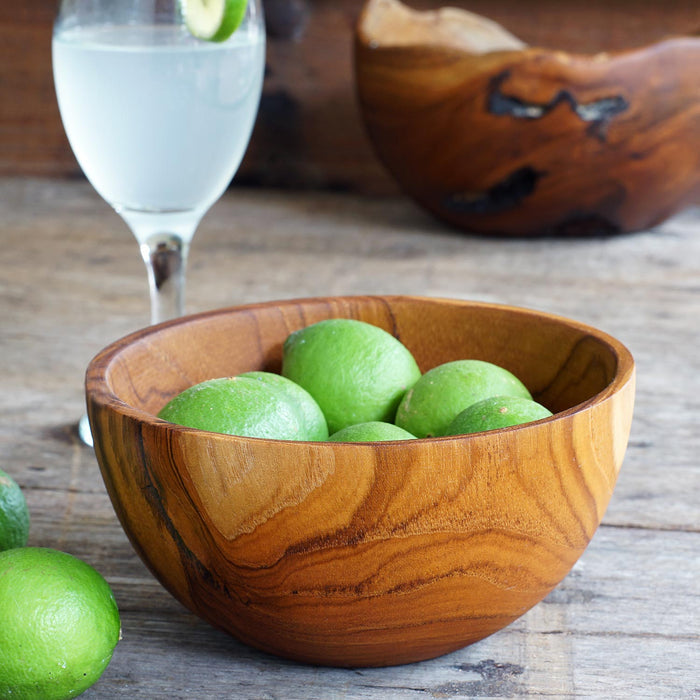 Rico & Plato chico teak bowl with limes inside next to 2 limes and a tall wine glass with a clouding white drink and lime slice on the rim all displayed on a wooden surface