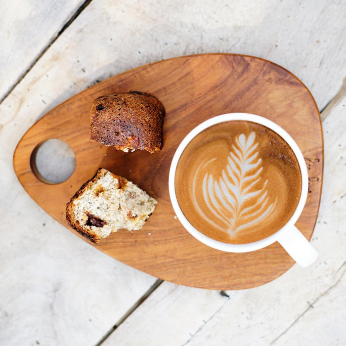 A teardrop-shaped teak wood coffee platter with a cup of coffee and a piece of bread on it, placed on a wooden surface.