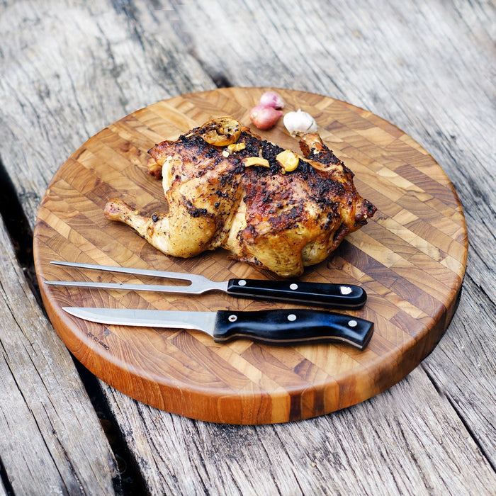 Circular end grain teak butcher block with knife, fork, and grilled chicken displayed on a wooden surface