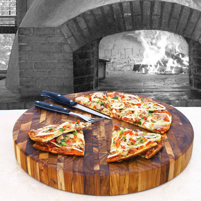 Circular end grain teak butcher block with knife, fork, and pizza slices on top placed on a white counter with fireplace in the background