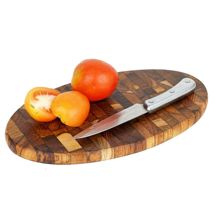 Oval end grain teak cutting board with tomatoes and knife on top displayed on a white background