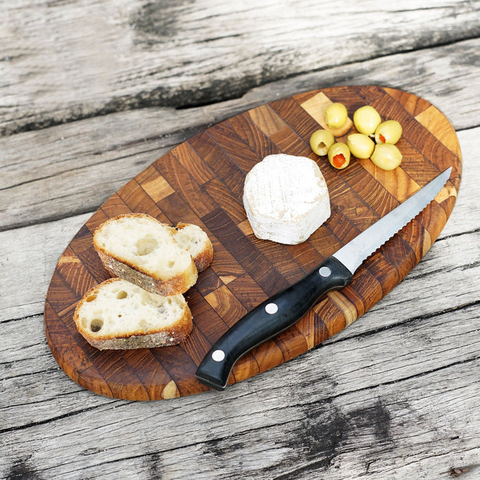 Top view of oval end grain teak cutting board  with olives, bread, cheese, and knife placed on top displayed on a wooden surface