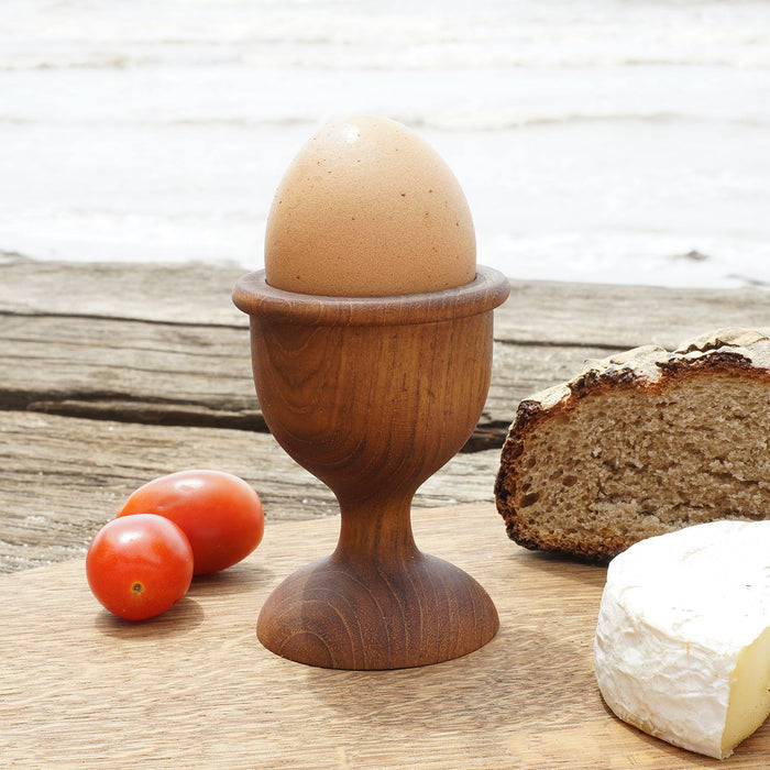 Teak egg cup with brown egg inside next to bread, cheese, and 2 tomatoes placed on a wooden surface outside