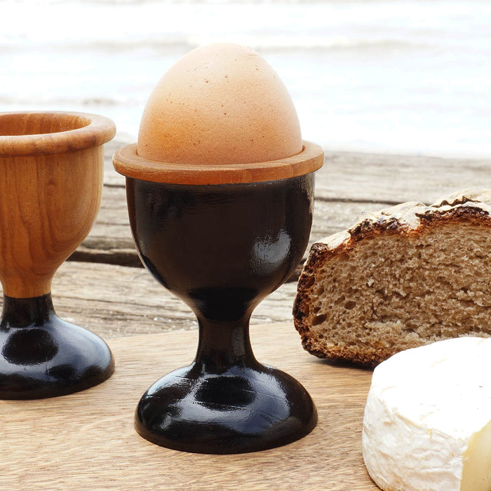 Two teak wood egg cups with one having a brown egg on it. One egg cup has a black-painted base and the other is natural teak with black colored foot.
