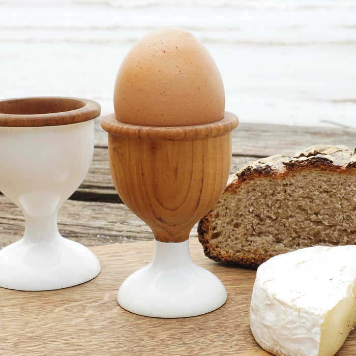 Two teak wood egg cups with one having a brown egg on it. One egg cup has a white-painted base and the other is natural teak with white colored foot.