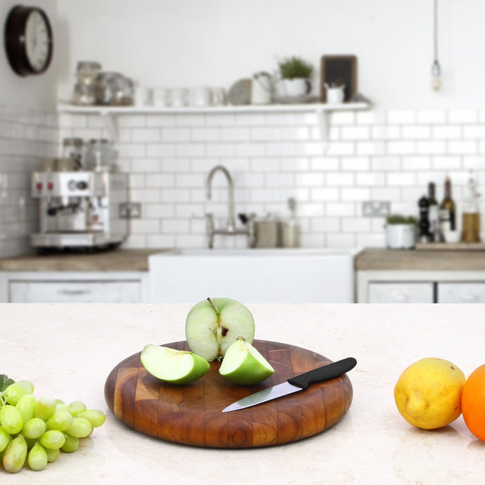 Circular end grain teak cutting board with sliced applies and knife on top next to grapes and lemons placed in a kitchen setting