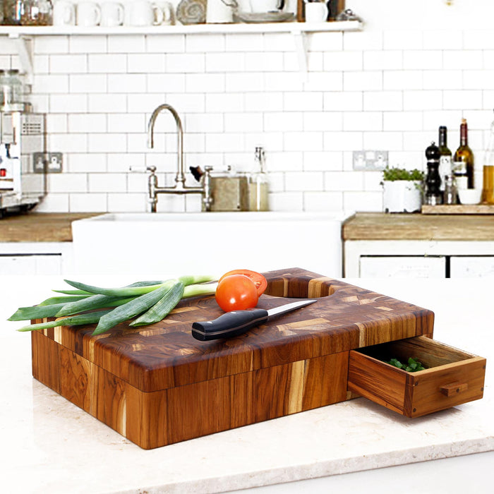 End grain teak chopping board with rubbish drawer open and hole on chopping board with tomatoes, knife, and green onions on top displayed in a kitchen setting