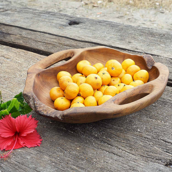 Rico & Plato dora teak serving bowl with integral handles with lemons inside bowl next to a flower displayed on a wooden surface