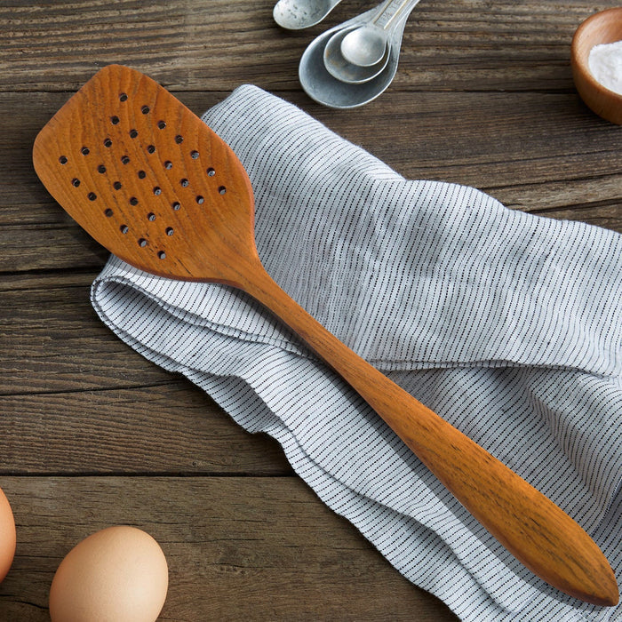 Rico & Plato emanuel teak spatula with air holes next to measuring spoons, bowl of salt, eggs, and laying on table cloth on a wooden surface