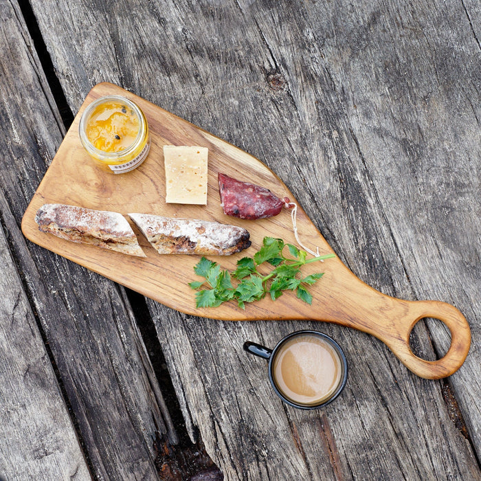 Rico & Plato epazote teak cutting board serving platter with jam, cheese, bread, herb, and cut up sausage link on top of platter next to a mug of coffee displayed on a wooden surface