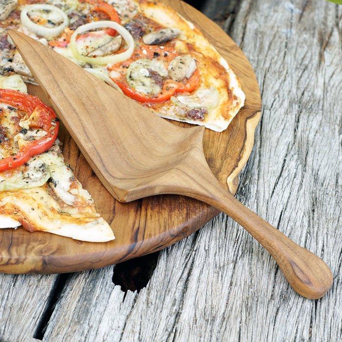 Teak pizza server sitting on a wooden board with pizza slices on top displayed on a wooden surface