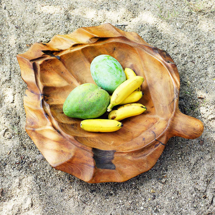 Top view of teak fruit bowl shaped as a leaf with fruit displayed on a dirt, gravel path