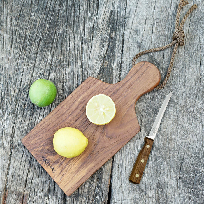 Rico & Plato ginger teak cutting board serving platter with a rope handle with a cut lemon and whole lemon on a platter next to a lime and knife displayed on a wooden surface