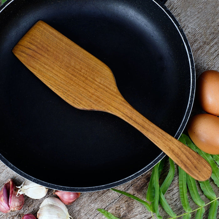 A teak spatula resting on a black frying pan, surrounded by eggs and herbs on a wooden surface