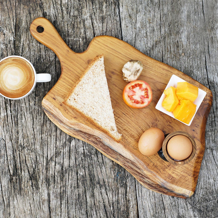 Top view of teak cutting & serving board with handle with bread, fruit, and eggs next to a creamy coffee displayed on a wooden surface
