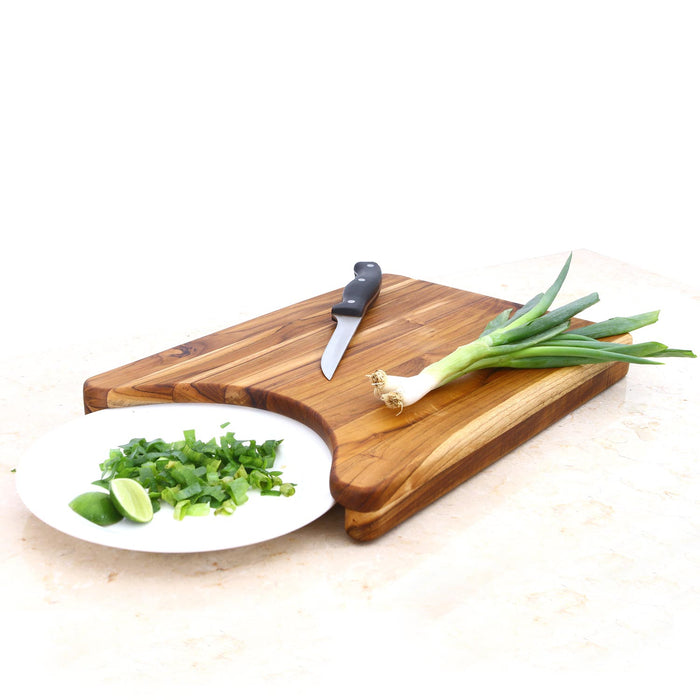 A rectangular teak wood chopping board with a curved cutout on the bottom. A knife and some green vegetables are on top of the board, with a plate of chopped green vegetables to the side.