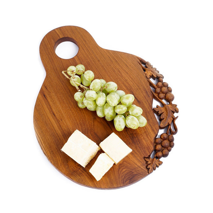 Teak cutting serving board with decoration carved on one side with a handle on the top and cheese blocks and grapes displayed on a white background