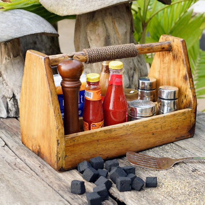 A rustic wooden BBQ caddy with a handle, filled with various condiment bottles and containers, placed on a wooden surface with a grill and some charcoal briquettes in the background.